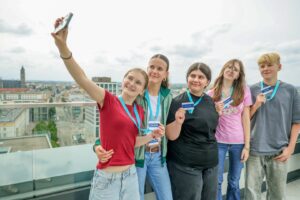 Five teens on a rooftop posing for a selfie, one arm raised with a phone, city skyline in the background, wearing blue lanyards with badges.