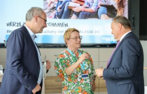 Three colleagues in business attire engage in a discussion in a bright room, with a large screen showing a welcome banner in the background.