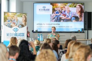 Speaker with microphone addressing seated audience at Medienklasse event; large screen and banner displaying students outdoors.
