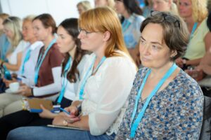 Group of professionals seated at a conference, wearing blue lanyards and taking notes.
