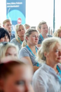 Audience at a conference or seminar, focusing on a woman with a blue lanyard in a striped shirt in the foreground.
