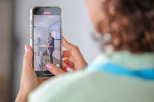 Person holds a smartphone filming a man in a suit giving a talk at a kitchen island showroom setup, seen on the phone screen.