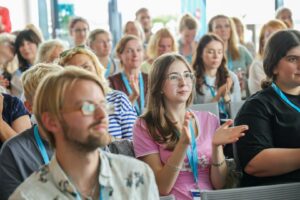 Front-row woman in a pink shirt claps while attending a conference with a diverse audience behind her.
