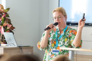 Female presenter with short blond hair wearing a colorful citrus-pattern shirt, speaking into a handheld microphone at a podium in a meeting room.