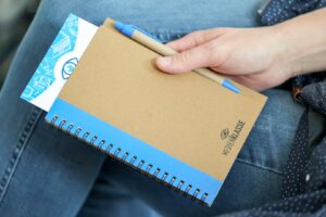 Person seated with a blue spiral notebook and a brown kraft notepad, holding a blue pen ready to write in a casual setting.