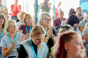 Audience of mostly adults seated and clapping at a conference or event, several wear lanyards and badges around their necks.