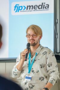 Man with a microphone speaking at a conference, wearing a floral shirt and blue lanyard, with a fip media banner in the background.