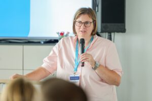 Speaker wearing a pink blouse and blue lanyard speaks into a handheld microphone at a conference.