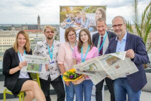 Group of six adults posing on a rooftop, two hold newspapers while a woman in pink holds a bouquet with a banner and cityscape in background