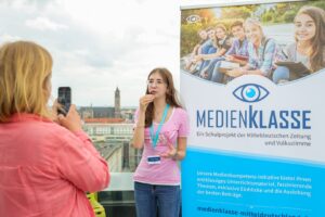 Young woman in a pink shirt speaks into a microphone while another person records her with a phone beside a Medienklasse banner outdoors on a rooftop city backdrop.