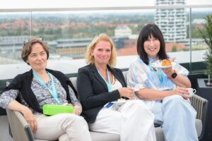 Three women on a rooftop terrace smile at the camera, with plates of cake and coffee in hand and a cityscape behind them.