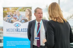 Man in a suit with a blue lanyard talks to a woman at a conference; a blue Medienklasse banner is in the background with an eye logo.