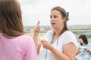 Woman in a white shirt with a blue lanyard speaks to another person on a rooftop, holding a small notebook and gesturing with her index finger.
