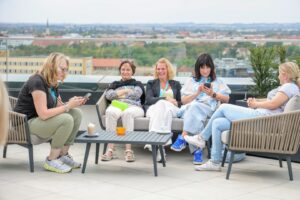 Five women sit on a rooftop lounge, chatting, with a distant city skyline in the background.