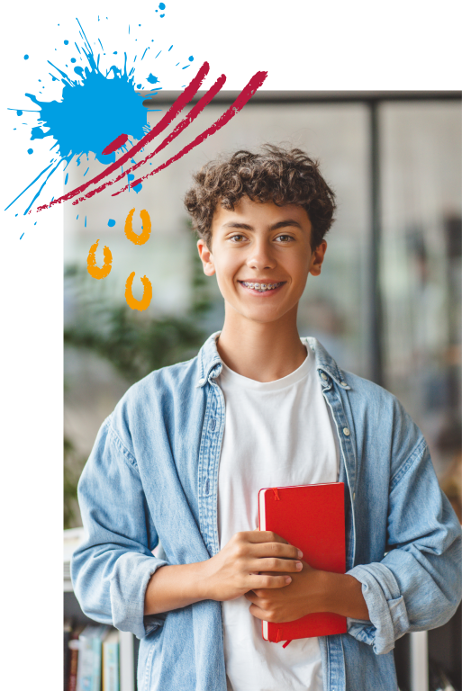 Smiling teenage boy with braces in a denim shirt holds a red notebook, decorative paint splashes overhead, indoor background behind glass doors.