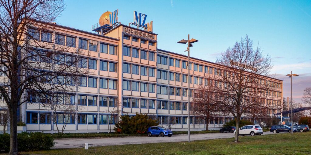 Office building with many windows and a Mediengruppe sign on the roof, blue sky and leafless trees in the foreground, cars parked along the street.